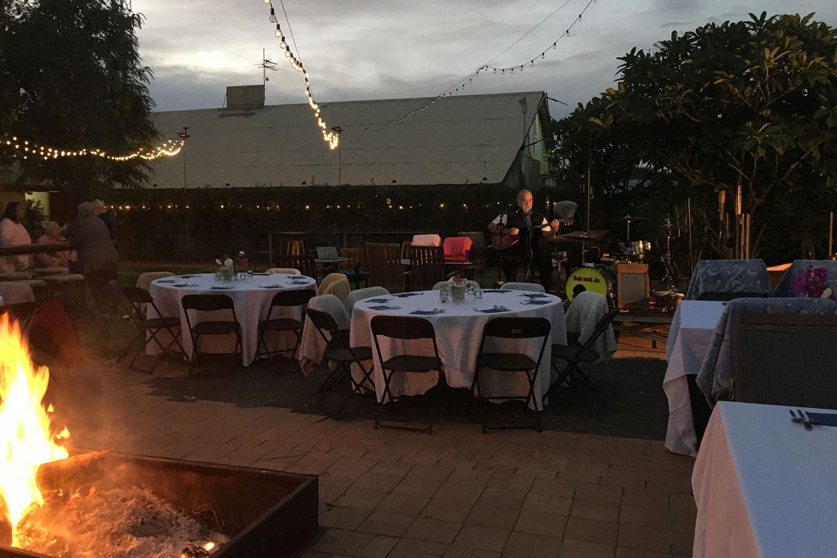 A fire pit burning beside white-clothed tables under festoon lights in the hotel garden at dusk, musicians on the verandah behind.