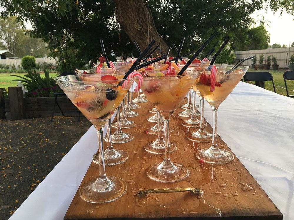Rows of cocktail glasses with fruit and striped straws lined up on a timber board on a garden table.
