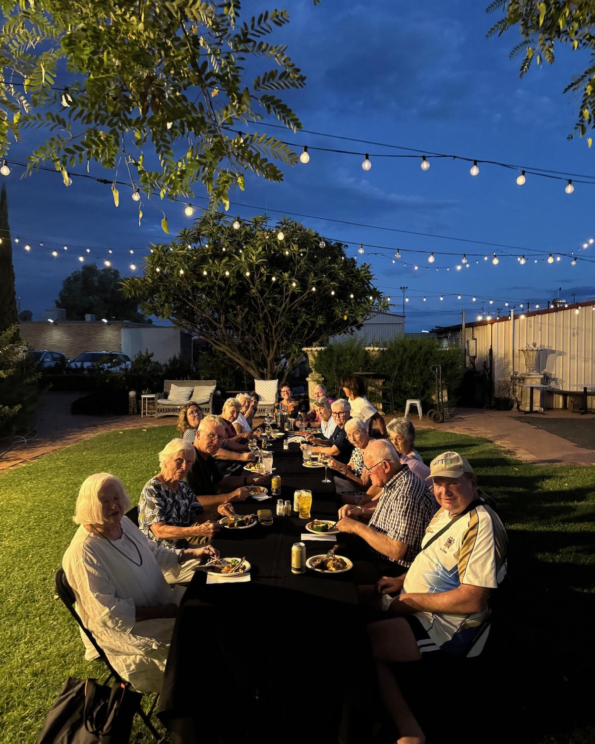 Guests at a long table on the Club Boutique Hotel lawn at blue hour, festoon lights strung through the trees above.