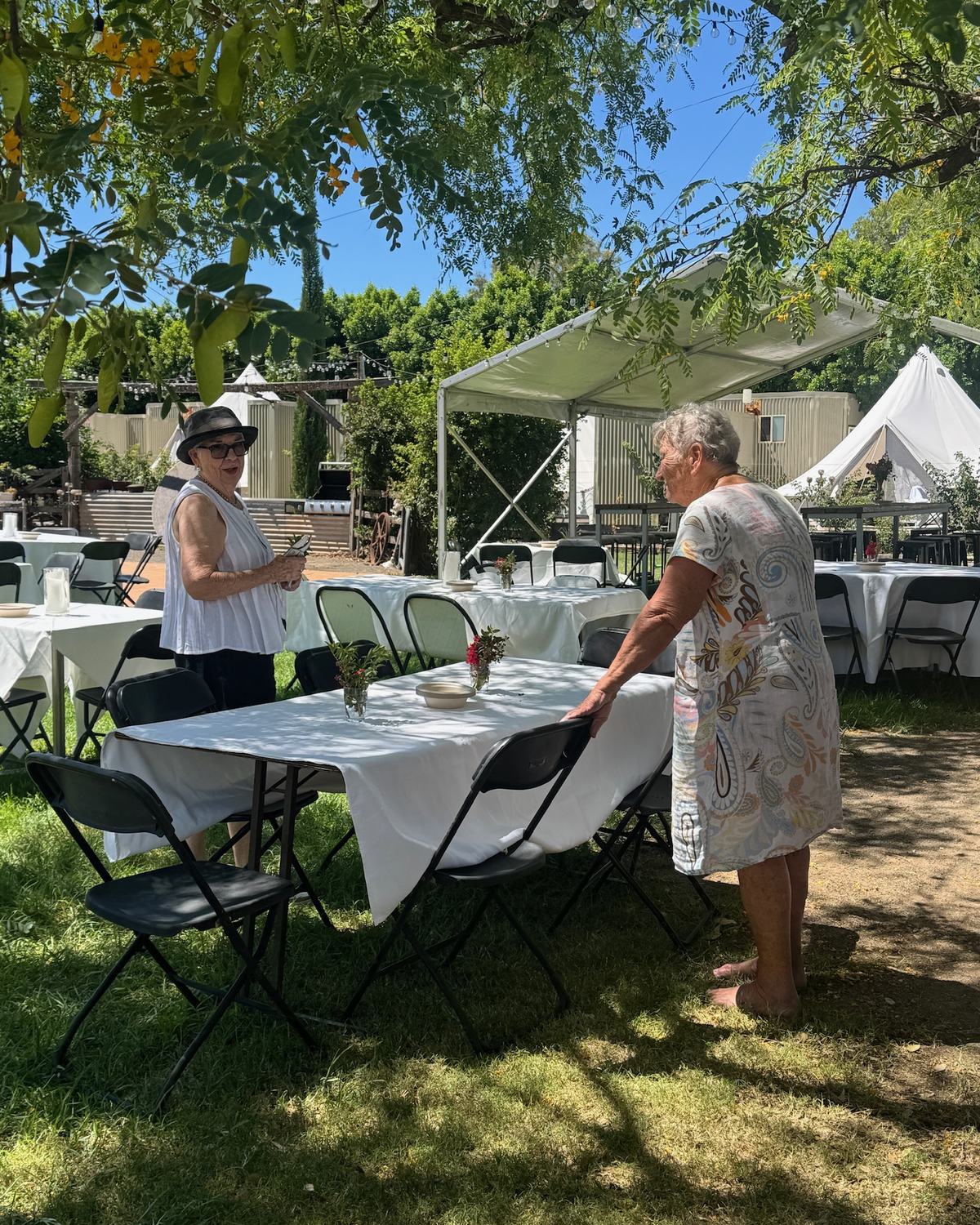 Tables with white cloths being set for lunch under the trees on the Club Boutique Hotel lawn, bell tents behind.