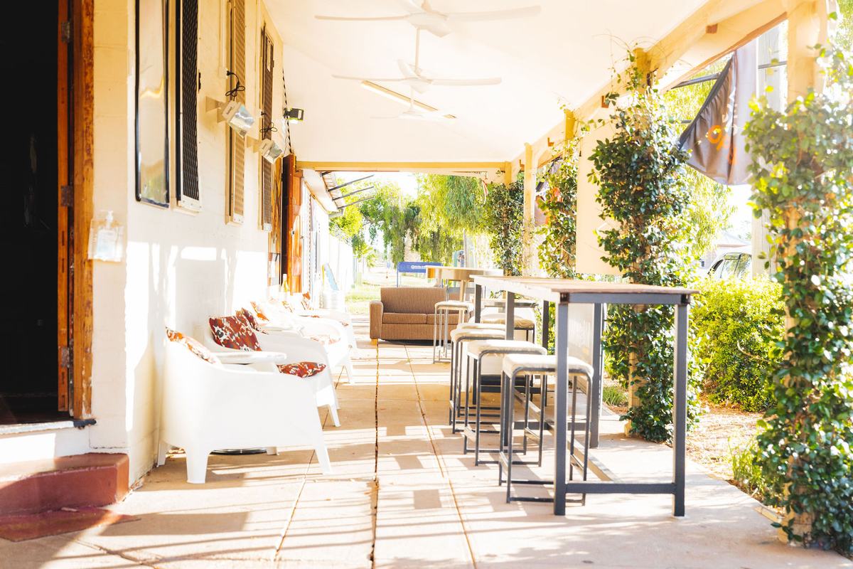 The long hotel verandah with white tub chairs, high tables, ceiling fans and vines in afternoon sun.
