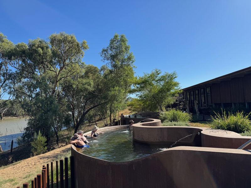 Bathers in sun hats in a curving soaking pool on the riverbank, gums and blue sky above.