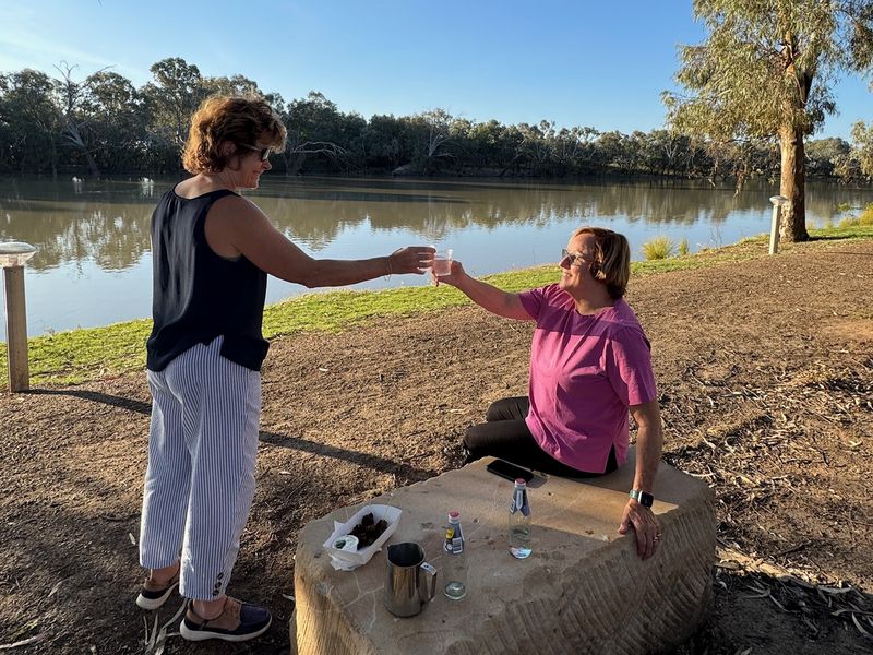 Two guests share a drink on a sandstone block beside the Warrego River in warm late-afternoon light.