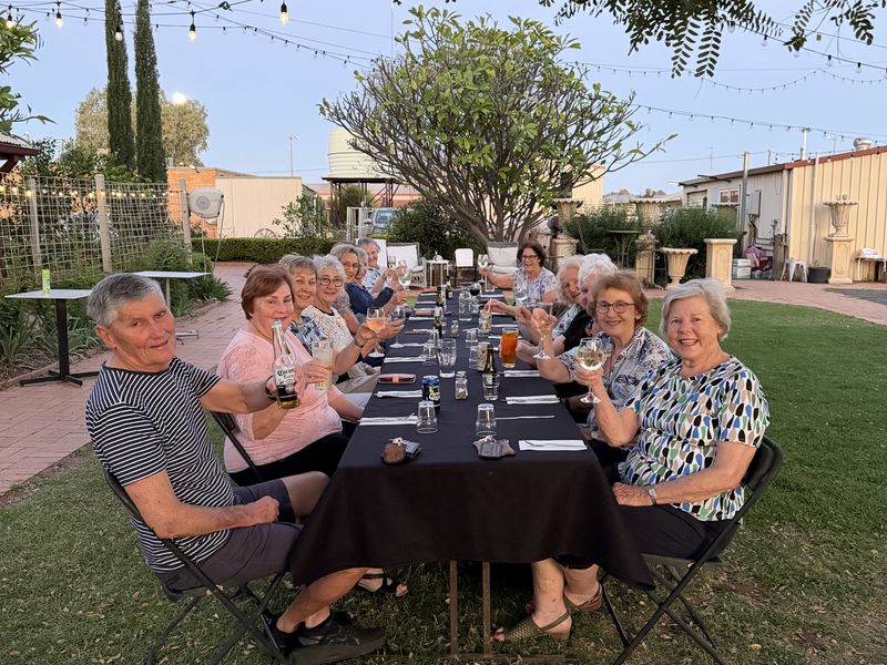 Guests raise their glasses along a long table on the hotel lawn at dusk, festoon lights strung through the trees overhead.