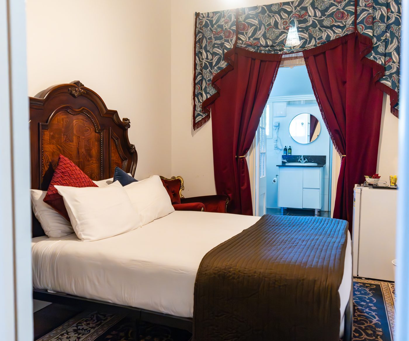 A heritage hotel suite with a carved timber bed and red velvet curtains framing the ensuite door.