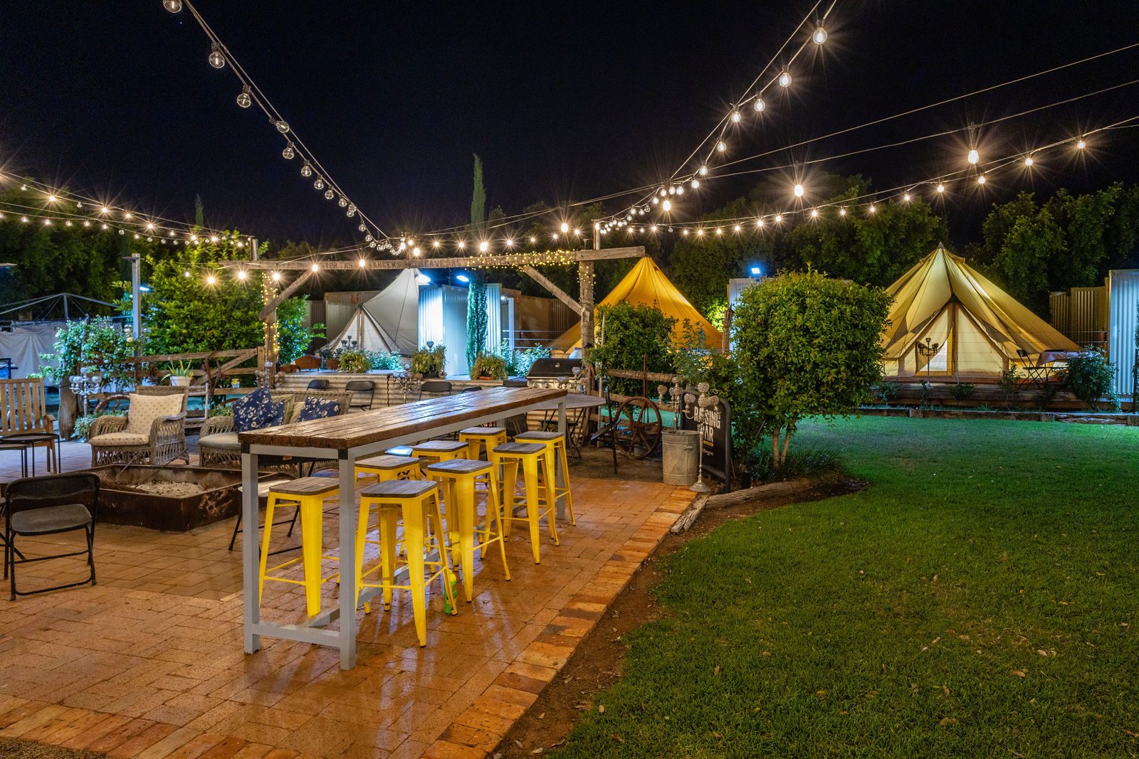 The hotel garden in the evening: a long table and yellow stools under strings of lights, bell tents on the lawn beyond.