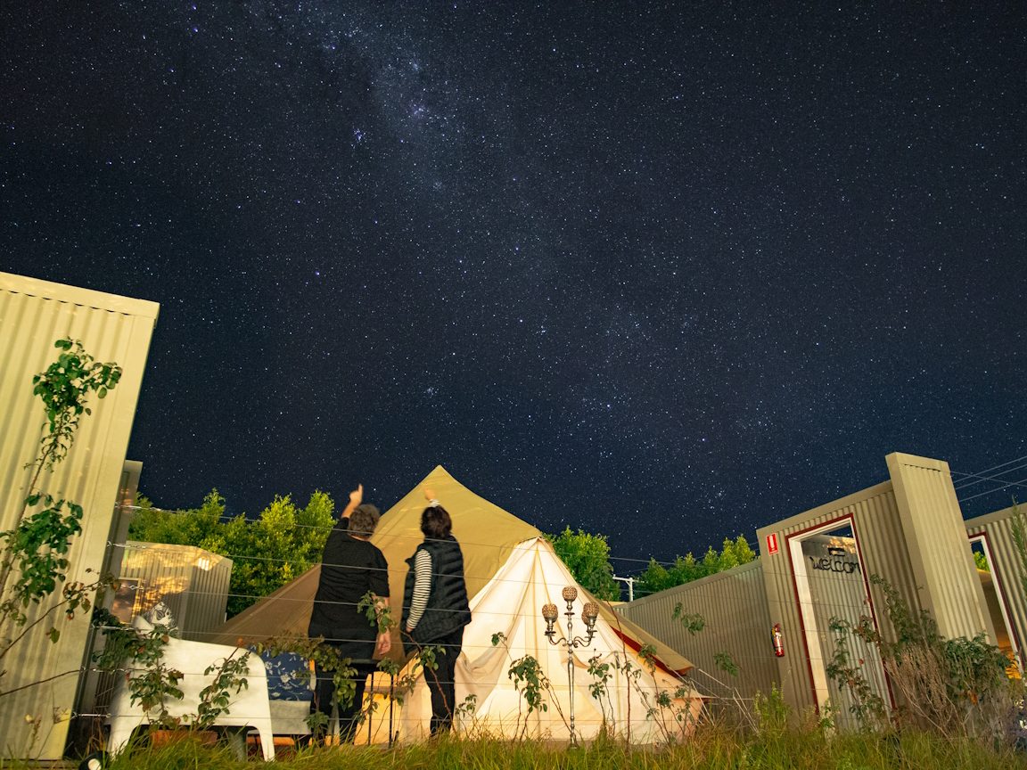 Two people stand on the garden lawn looking up at a sky full of stars, beside a glowing bell tent.