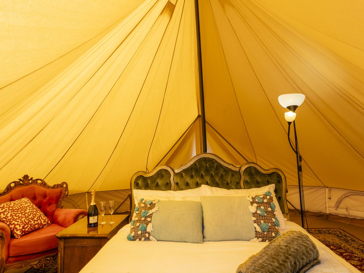 Inside a bell tent: a made bed with an ornate bedhead under the canvas, beside the centre pole and a reading lamp.