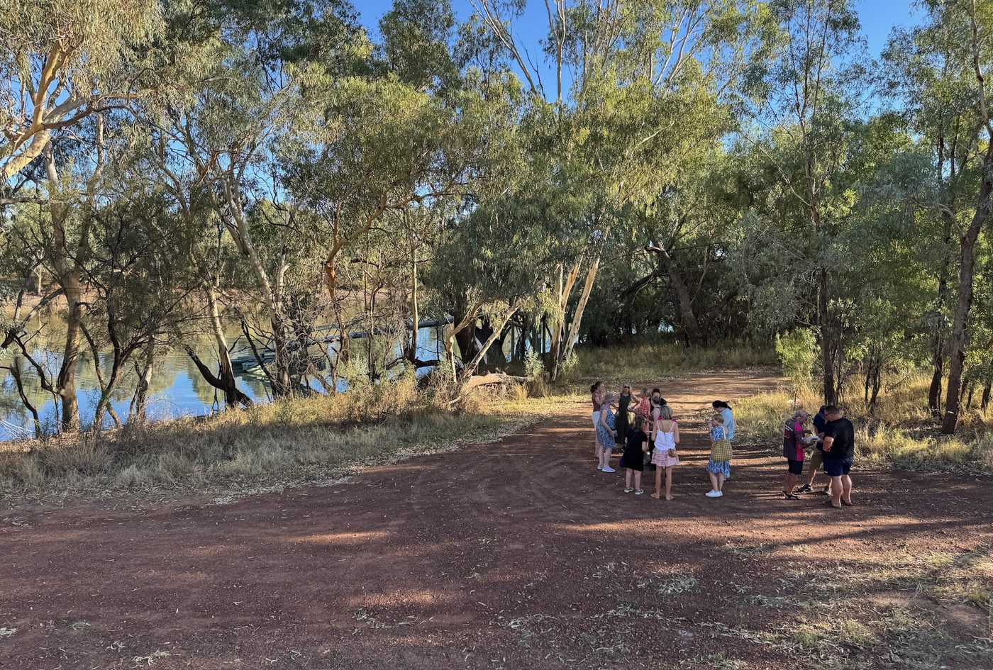 A small group gathers on a red dirt track beside the river, under tall gums.
